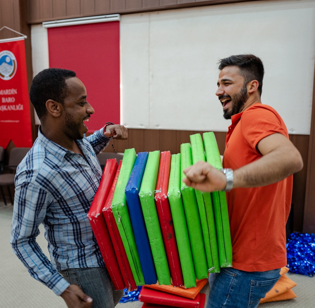 Two people balancing a stack of flat square pads between their stomachs