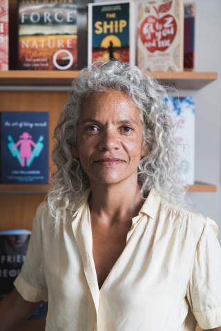 Image of the author Catherine Johnson. She is standing in front of a bookshelf and has shoulder length curly silver hair.